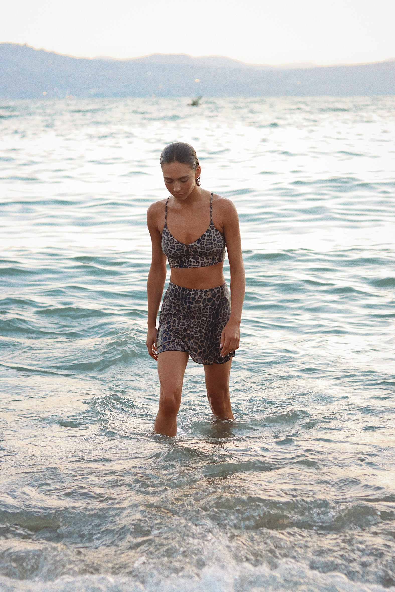 Woman in a leopard print bikini standing in shallow water with mountains in the background